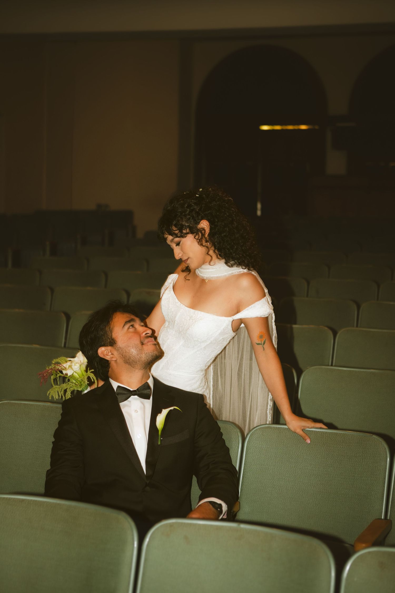 A bride in a white off-shoulder gown and groom in a tuxedo share a romantic moment in a vintage theater.