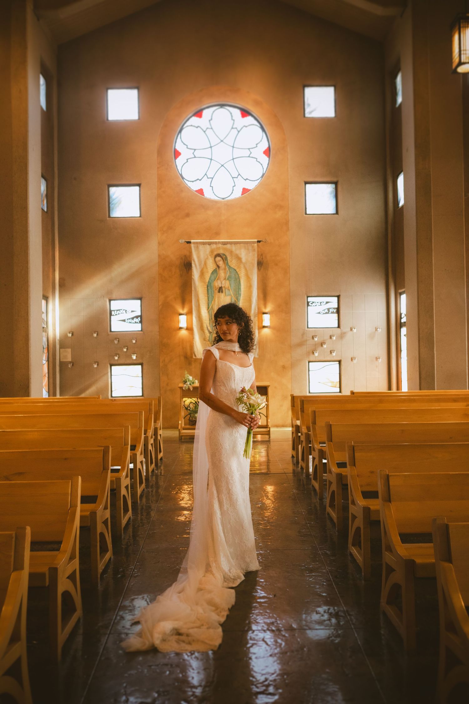 Bride in white gown stands in sunlit chapel aisle with stained glass rose window and Our Lady of Guadalupe banner.