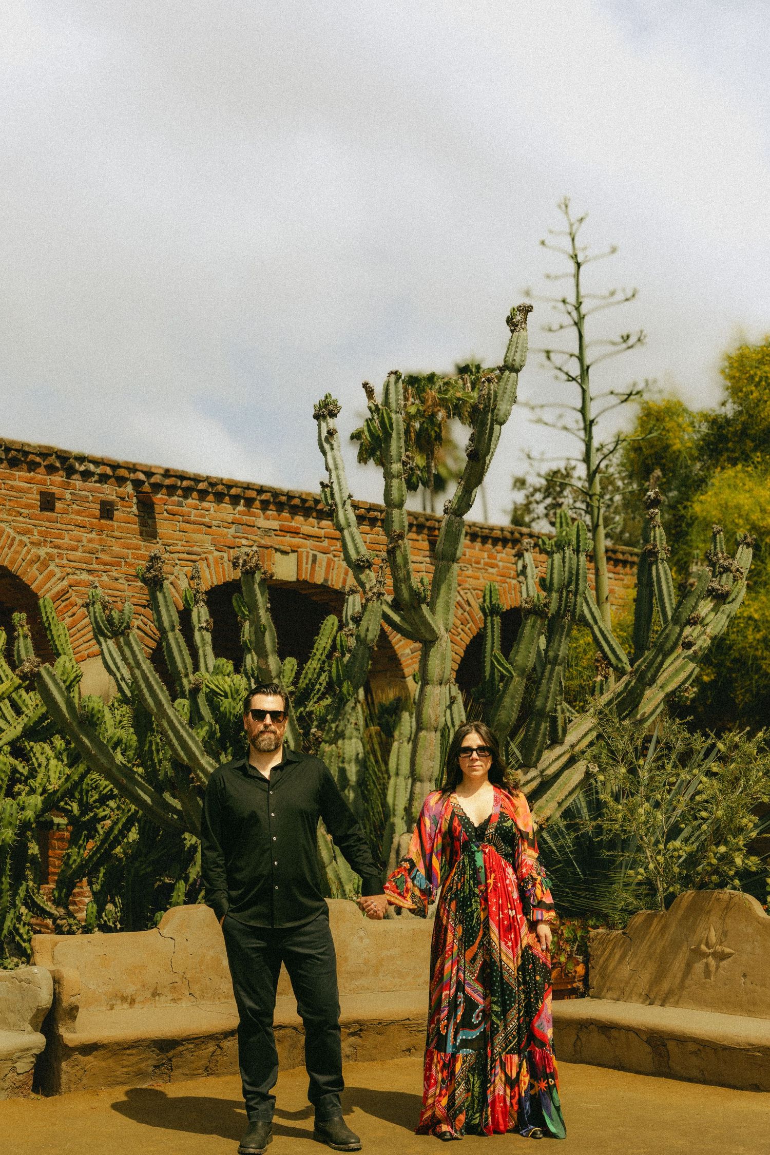 Couple holds hands before towering cacti at historic Spanish mission with brick arches in background.