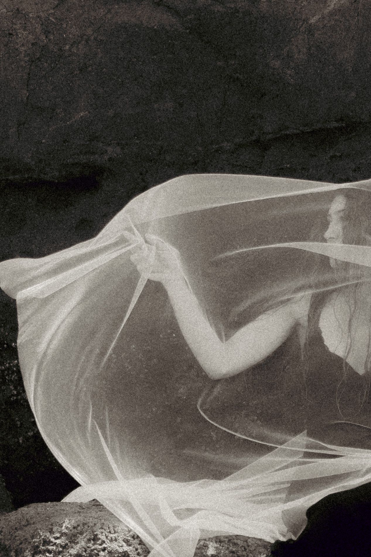 Black and white photo of a figure draped in flowing sheer white fabric against dark rocky background.