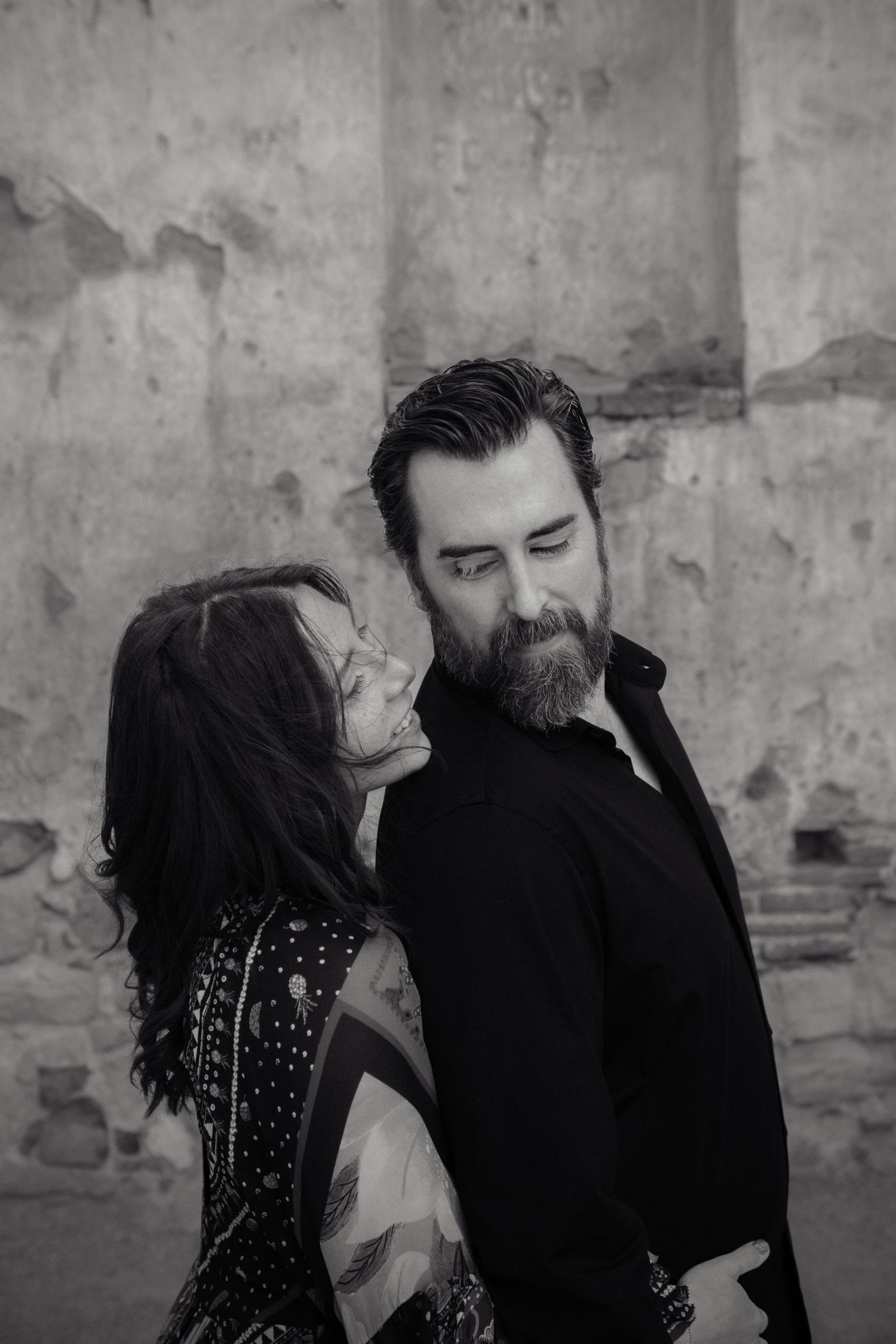 Black and white photo of a bearded man in black jacket with a woman leaning in close, posing against a textured wall.