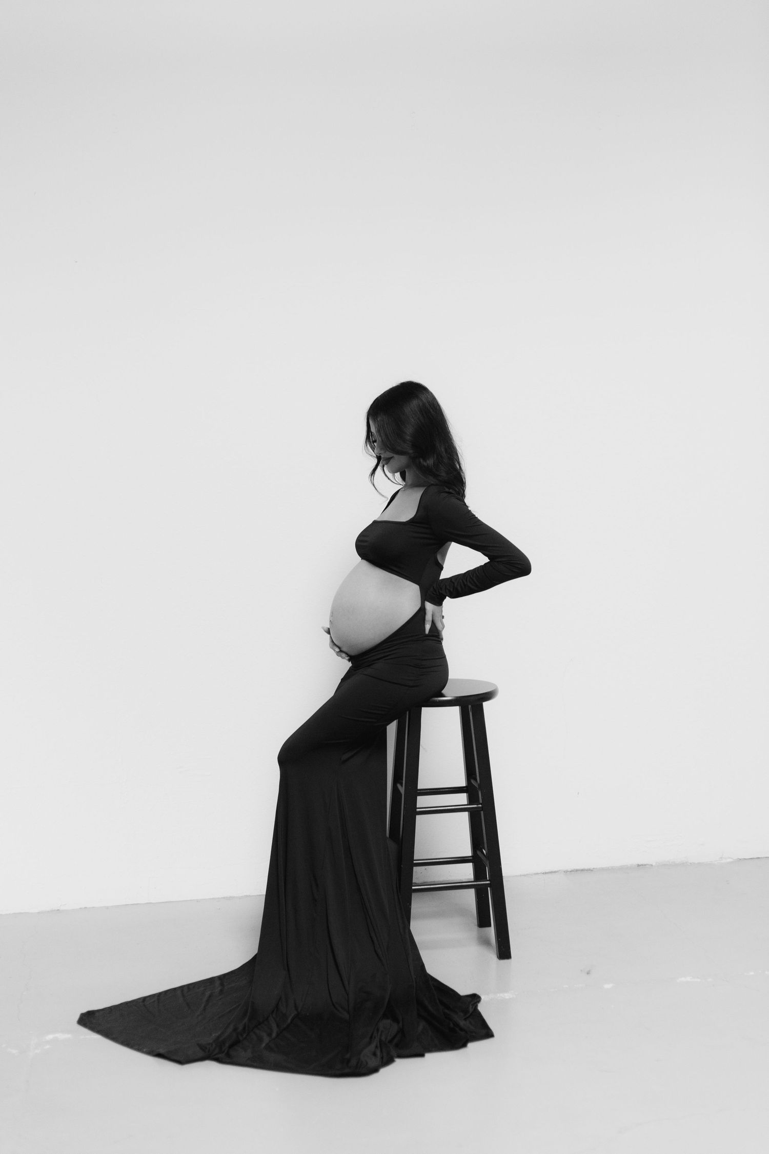 Elegant black and white studio portrait of pregnant woman in black gown seated on stool.