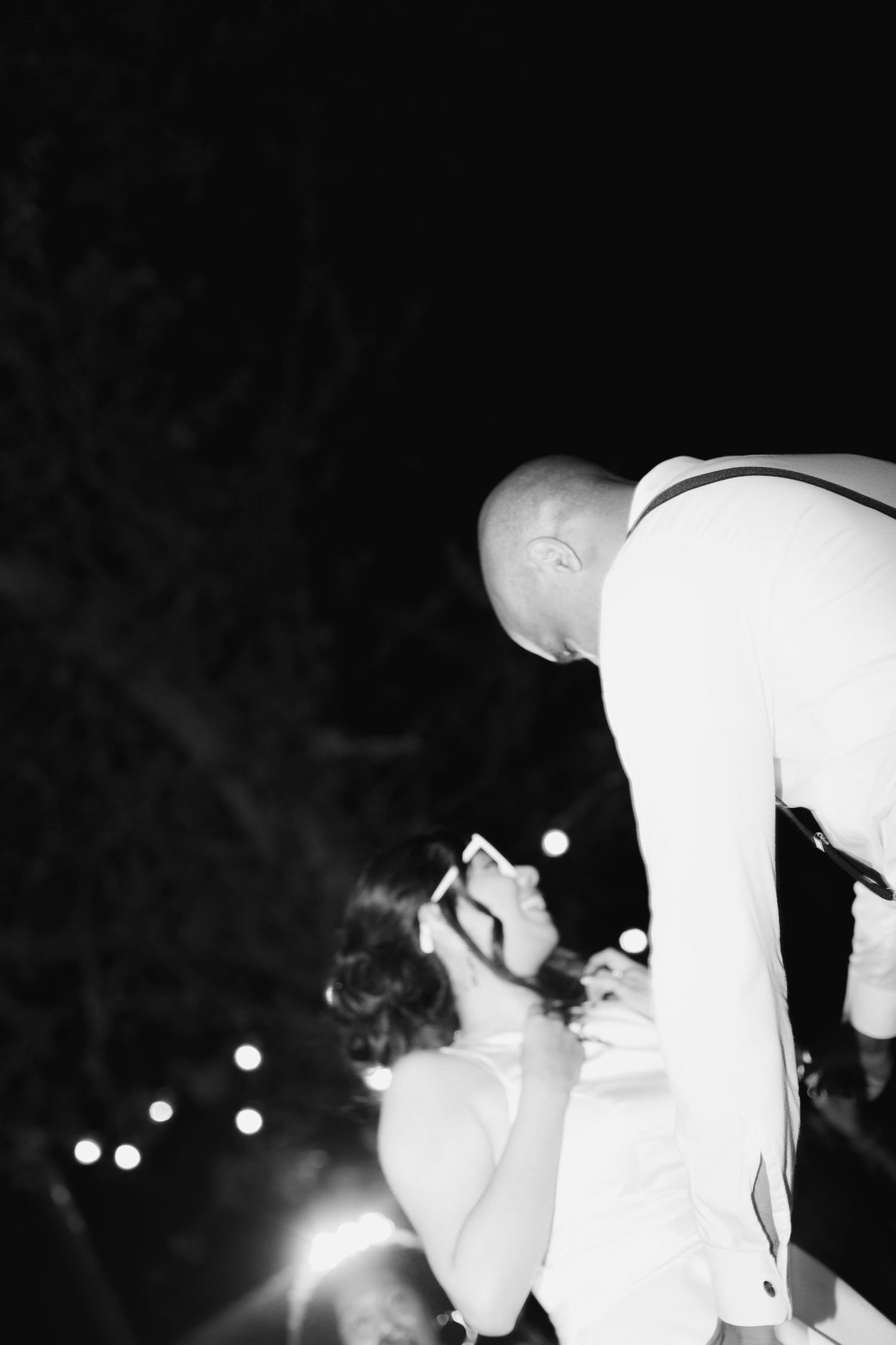 A bald groom leans toward his bride during an outdoor evening wedding reception with string lights.