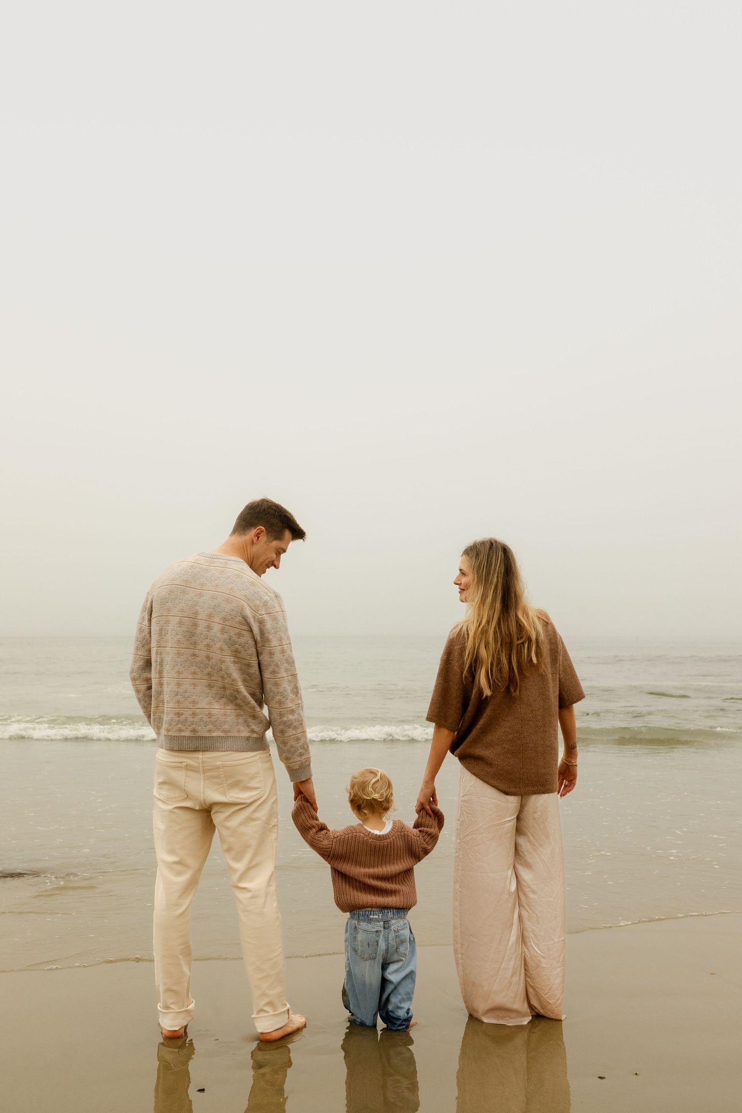 Family of three holding hands on sandy beach, parents looking down at young child between them.