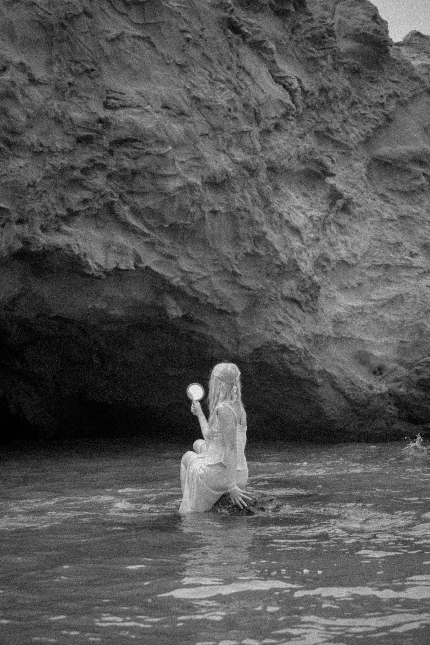 Ethereal woman in white dress standing in ocean water holding glowing orb near rocky cliffs, black and white.