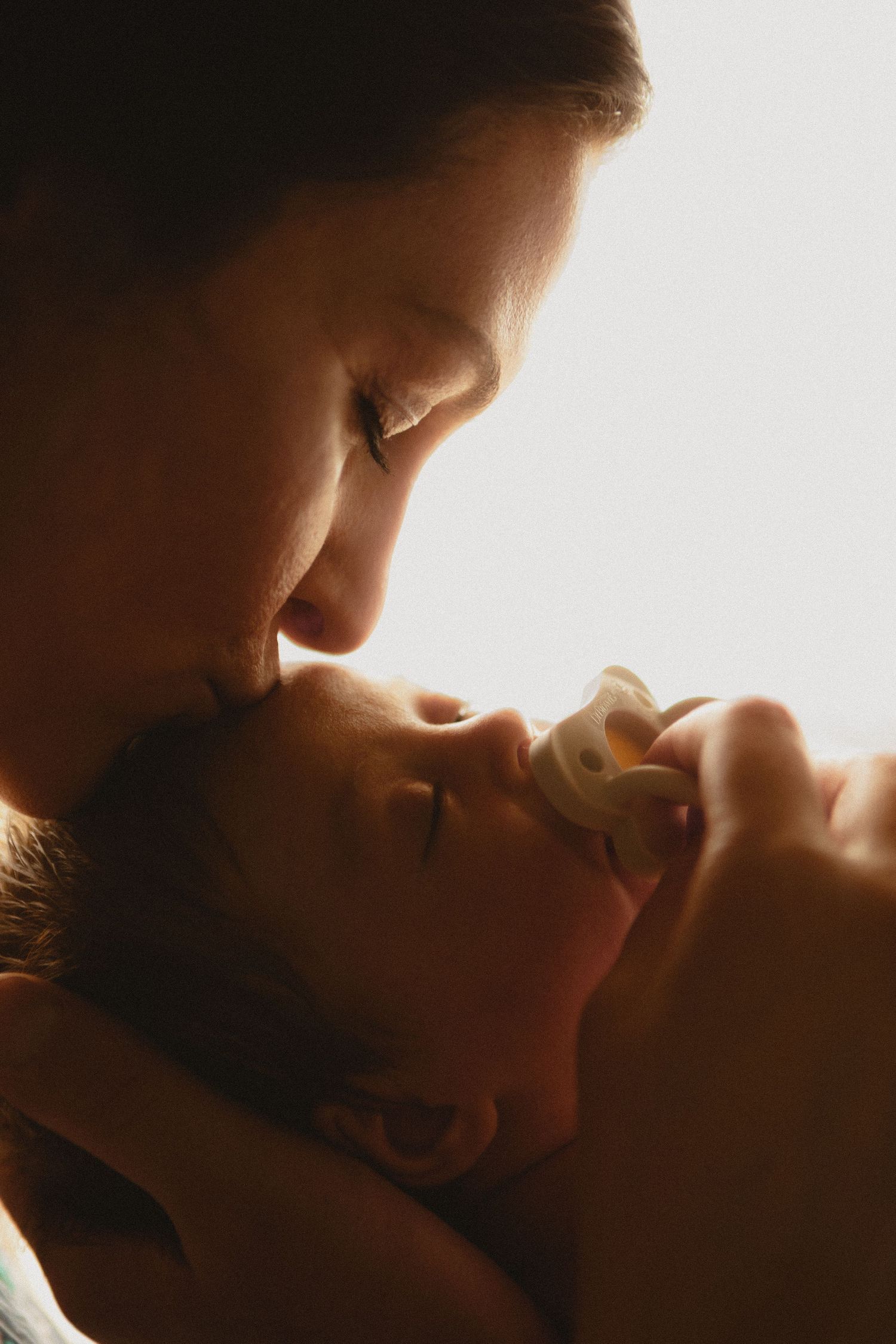 Mother tenderly kissing sleeping newborn baby in beautiful backlit  portrait.