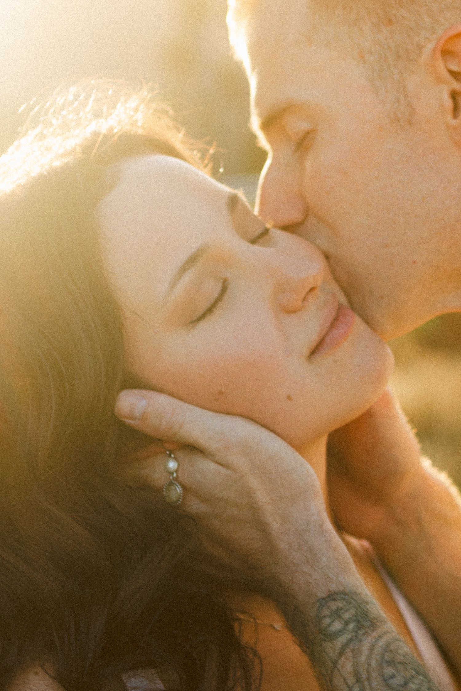 Man tenderly kissing womans temple while cradling her face in golden sunset light.