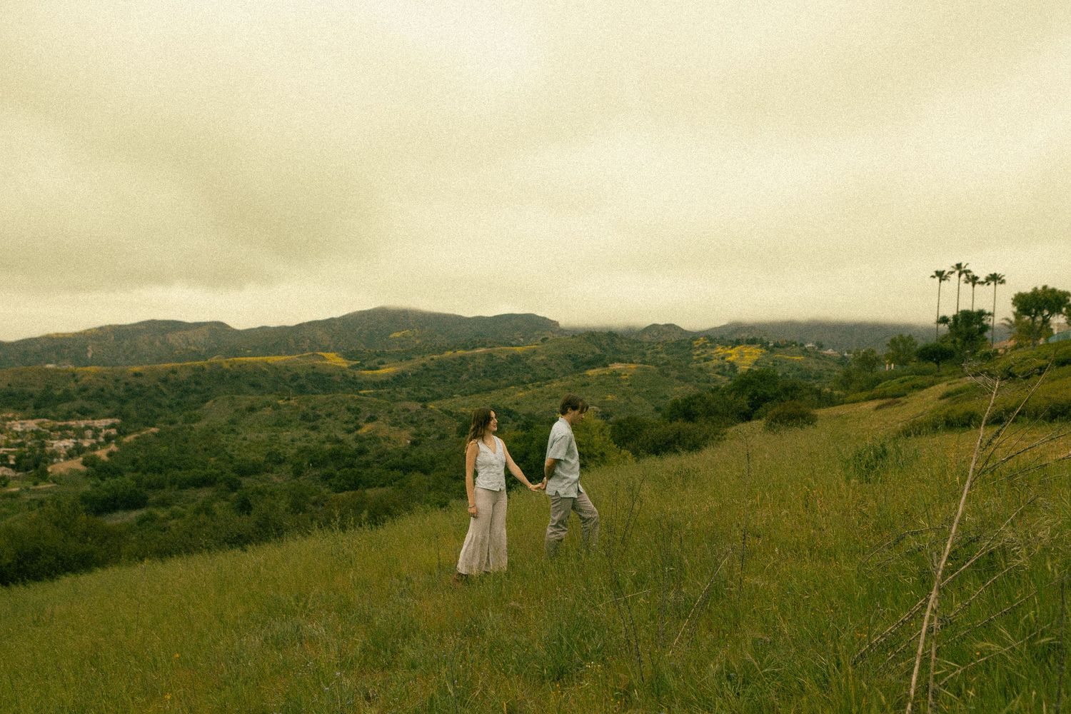 Couple walking hand in hand through green meadow with rolling hills and cloudy sky.