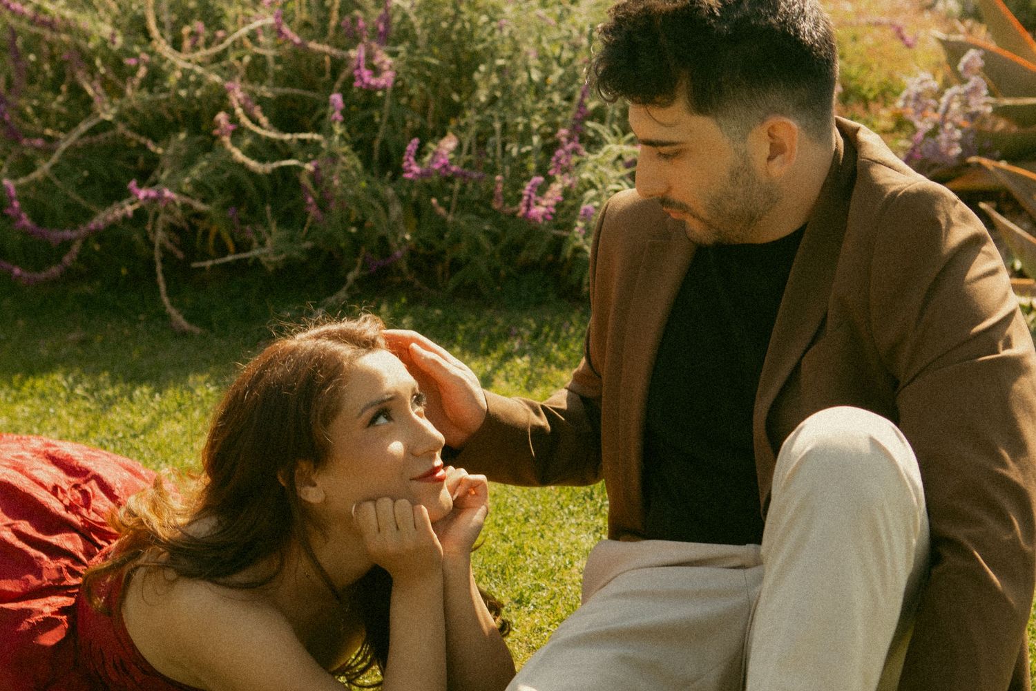Young couple shares tender moment on grass, woman in red lying down gazing up at man in tan blazer.