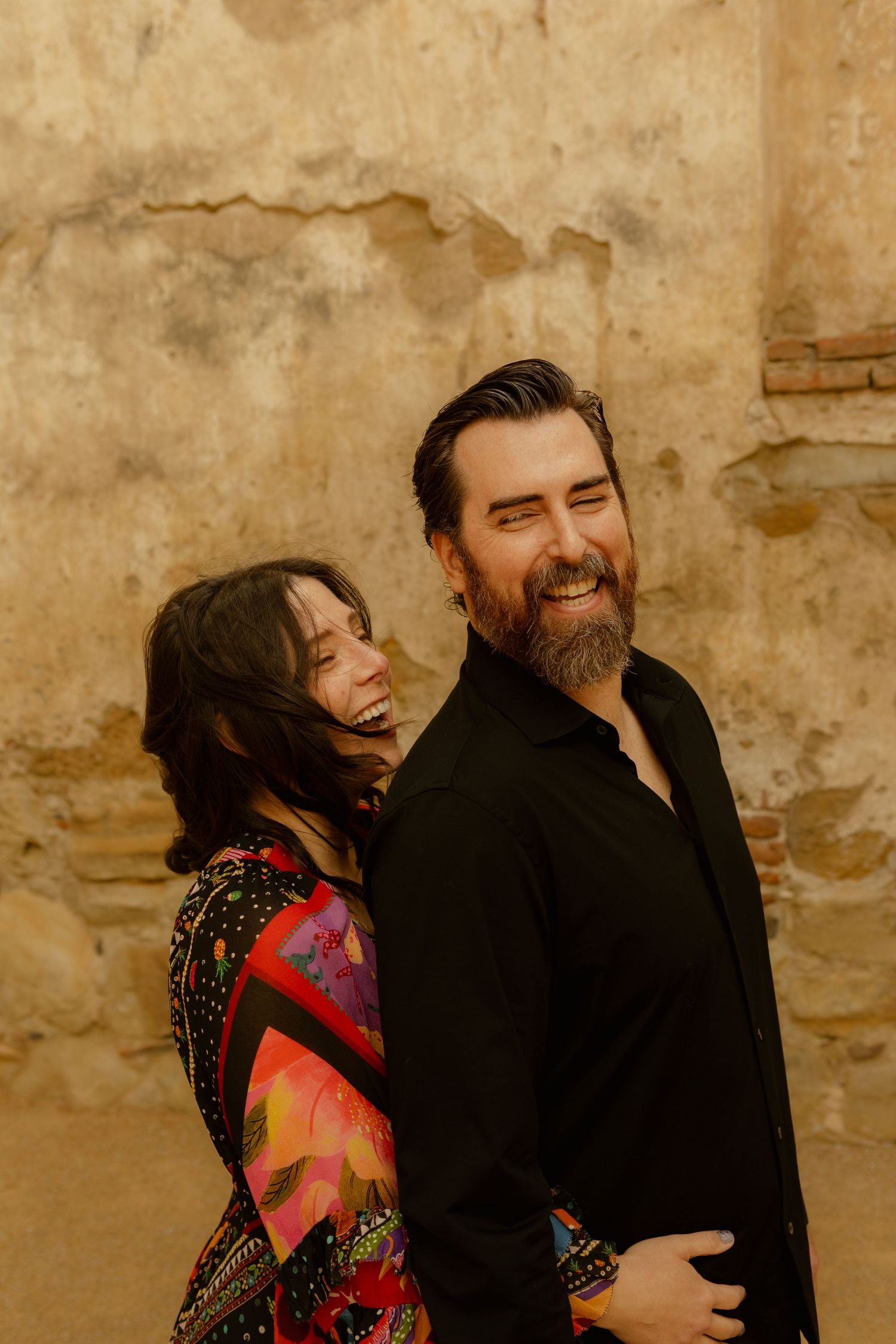 Laughing couple poses against rustic stone wall, woman in colorful dress leaning on bearded man in black shirt.
