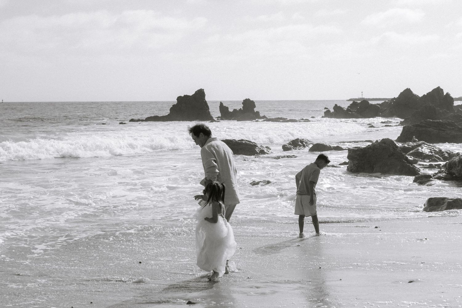 Black and white photo of a couple in wedding attire walking along a rocky beach shoreline with ocean waves.