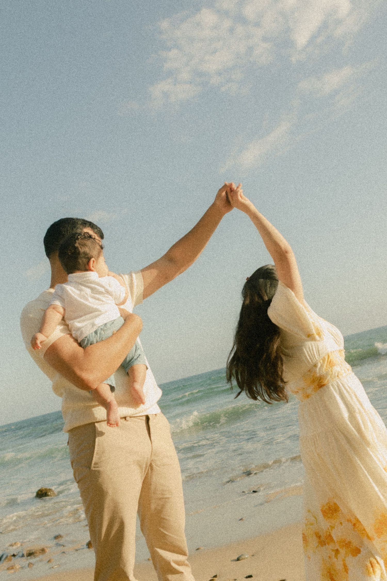 A couple dances on a sunny beach, man holding a baby, woman in floral dress, ocean waves in background.