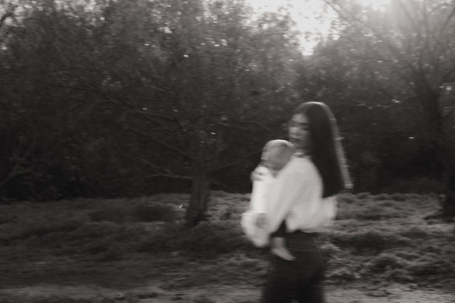 Black and white motion-blurred photo of a mother holding her newborn baby while walking outdoors.