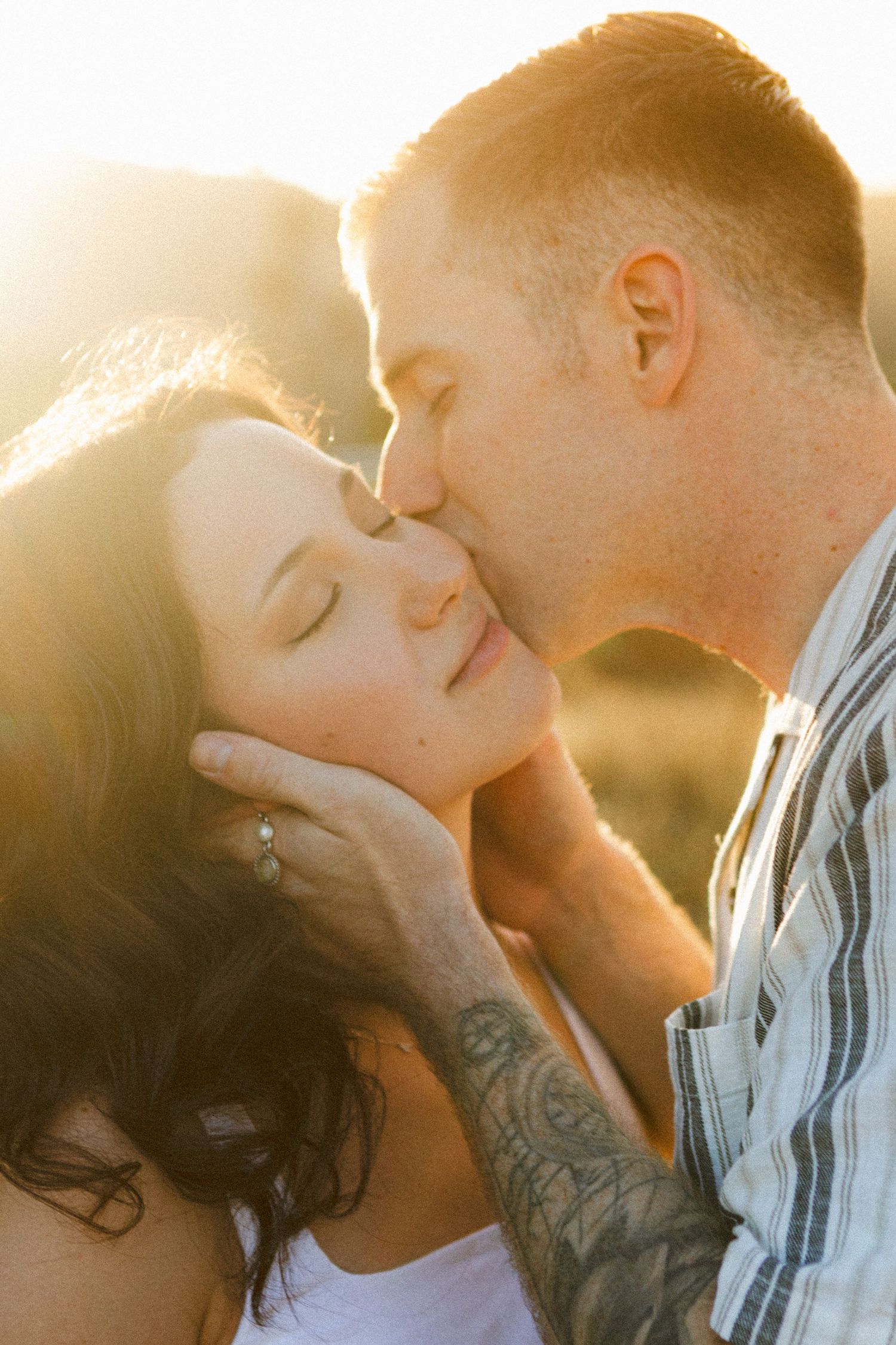 Tattooed groom kisses bride on cheek during golden hour shoot.