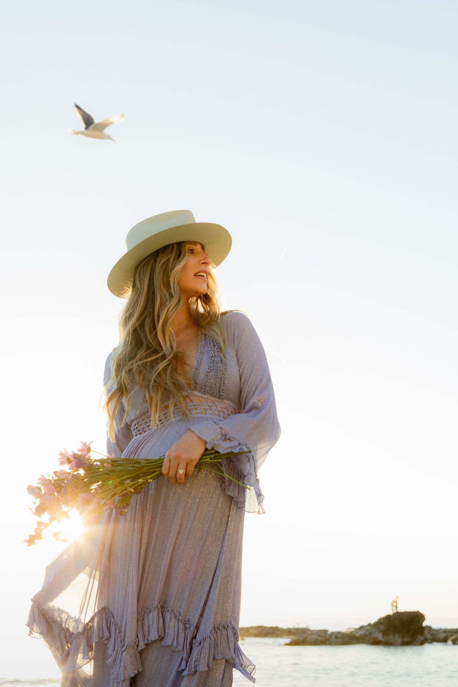 Bohemian woman in wide-brim hat and flowing gray dress holds pink flowers at golden hour beach.