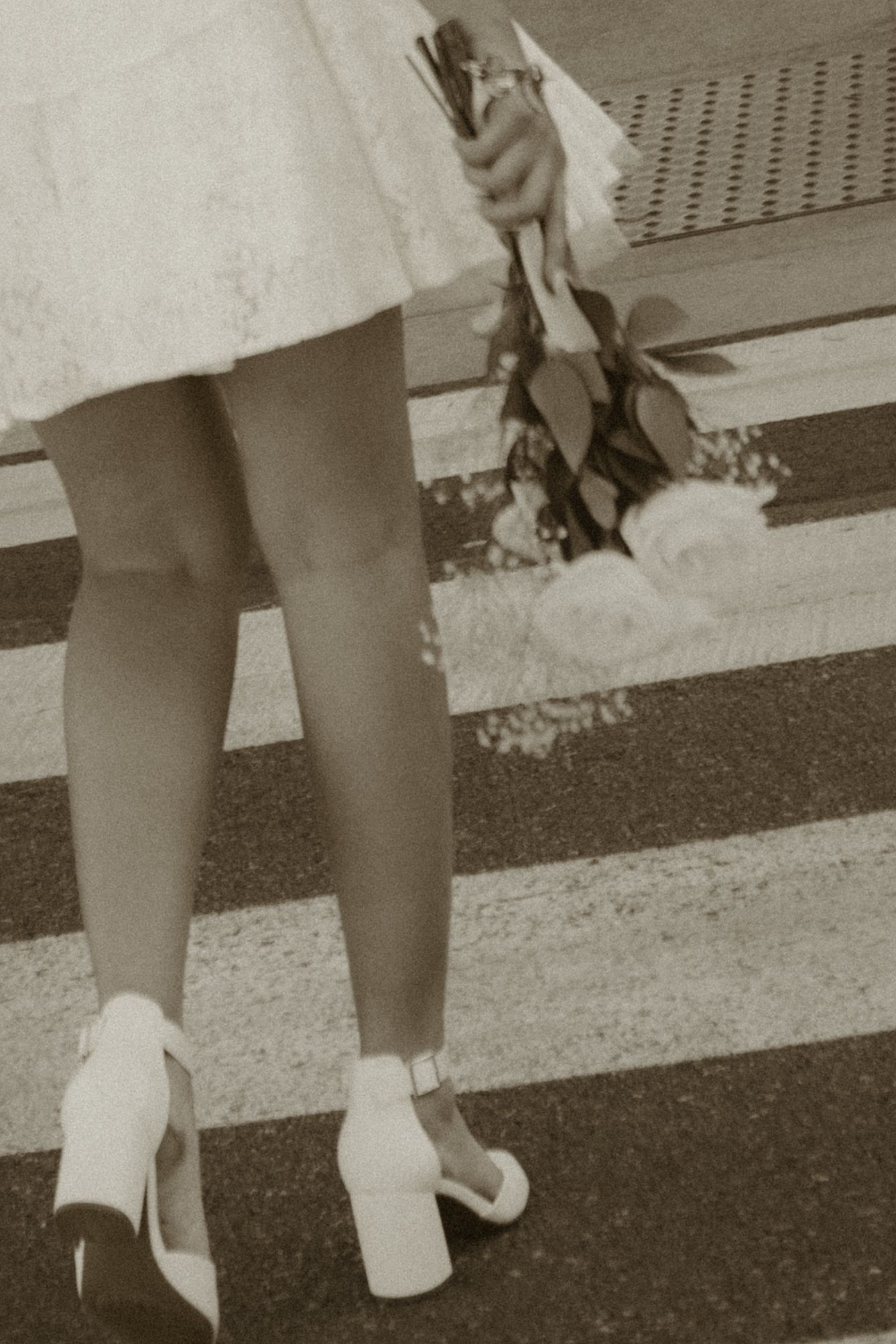 Bride in white lace dress and platform heels crossing zebra crosswalk with bouquet.