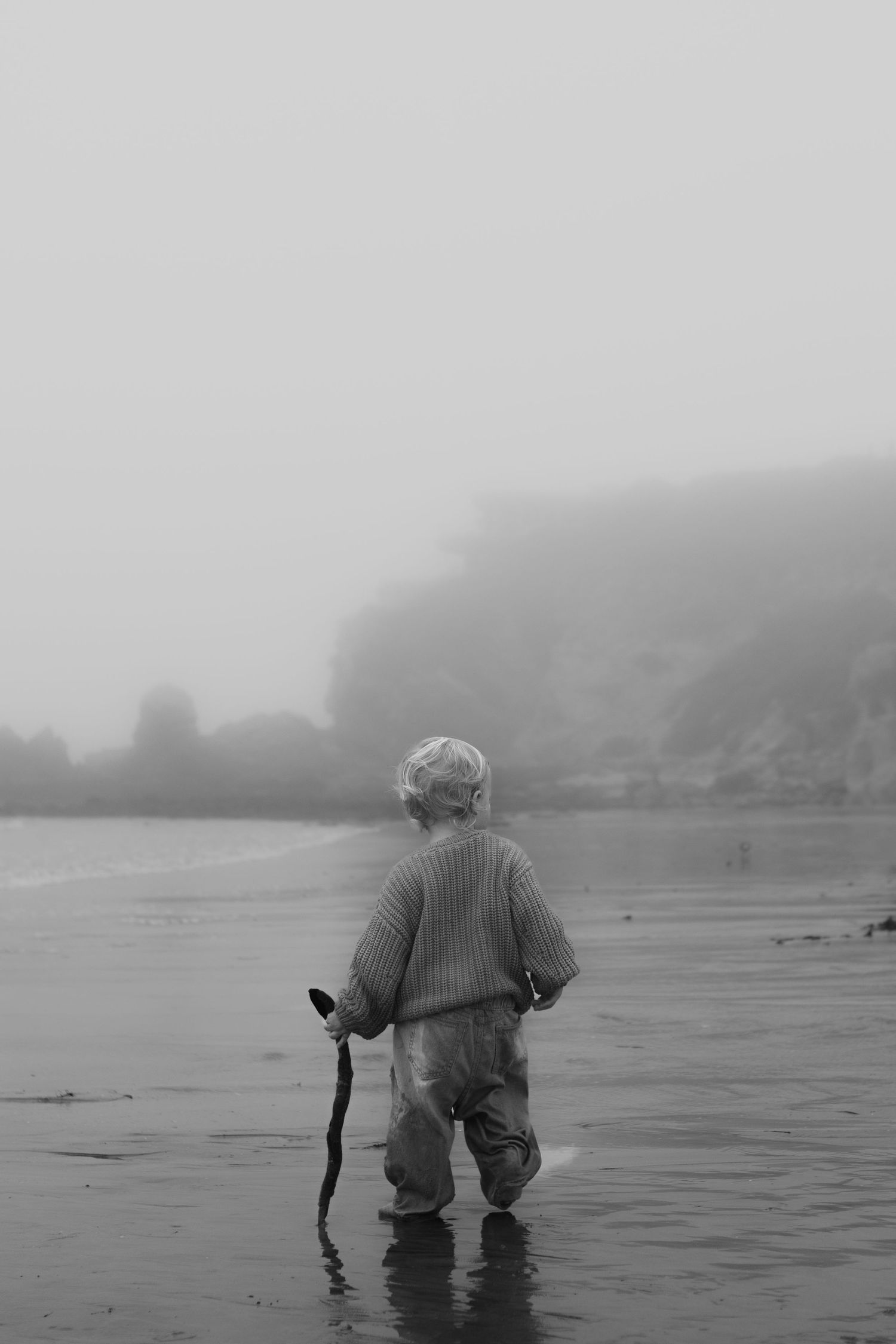 Young toddler standing alone on misty foggy beach holding a stick, black and white photo.