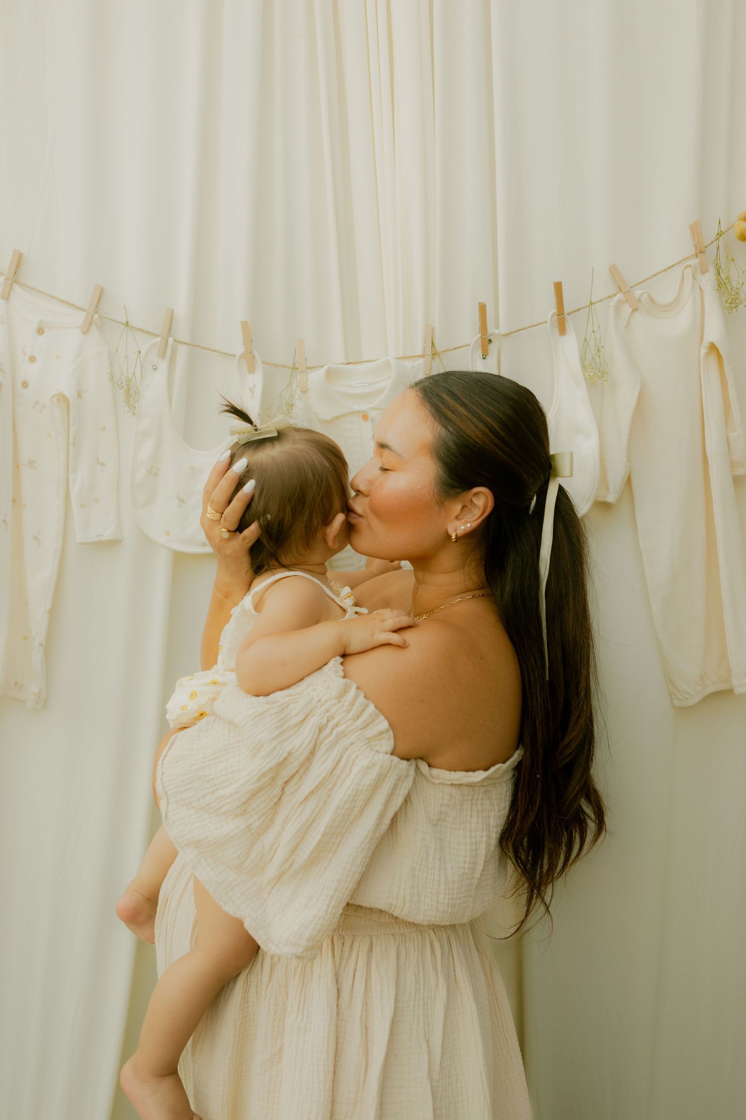 Mother in off-shoulder white dress kissing baby girl in front of hanging baby clothes.