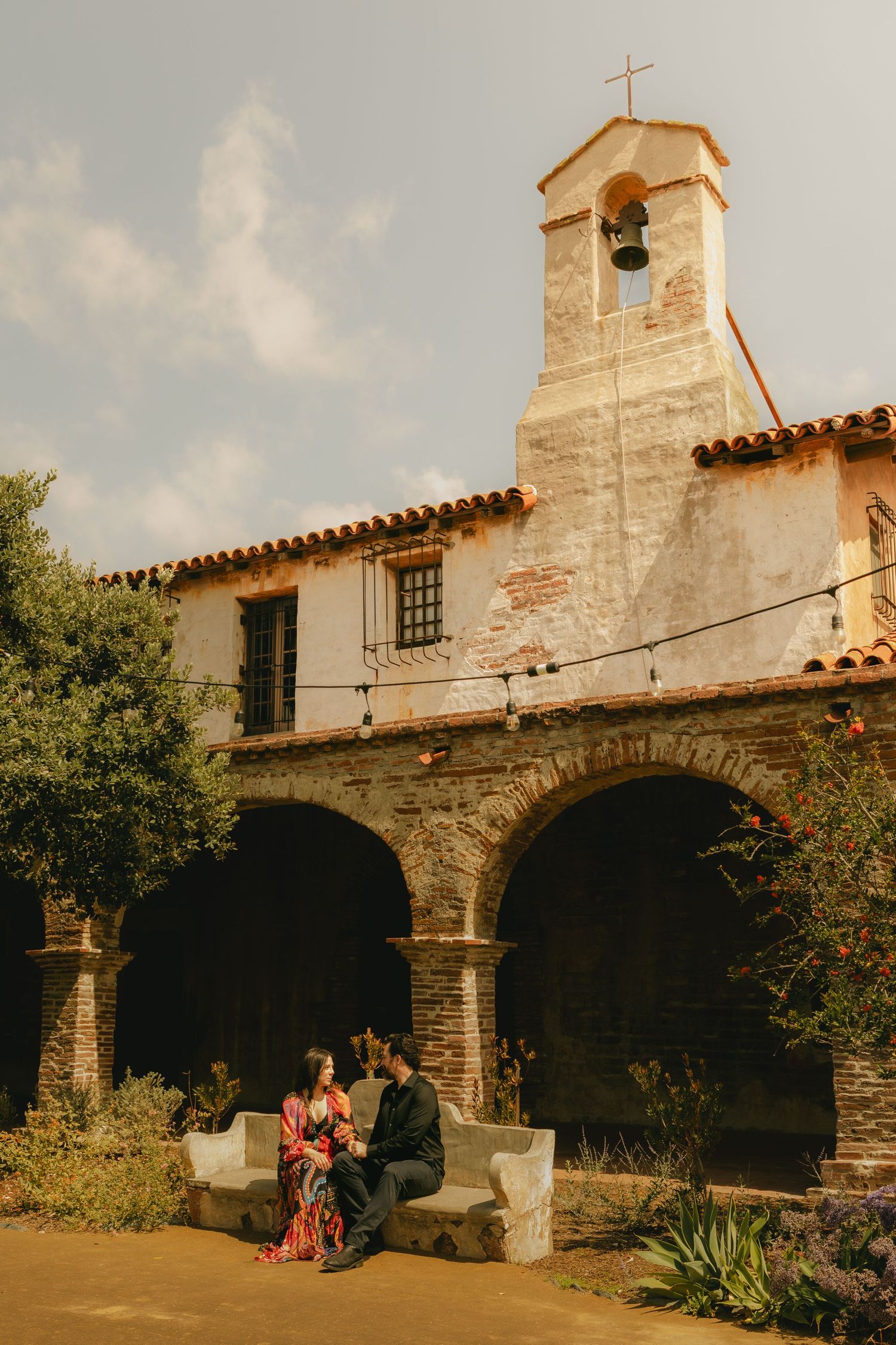 Couple sits on stone bench in courtyard of historic Spanish mission with bell tower and arched colonnade.