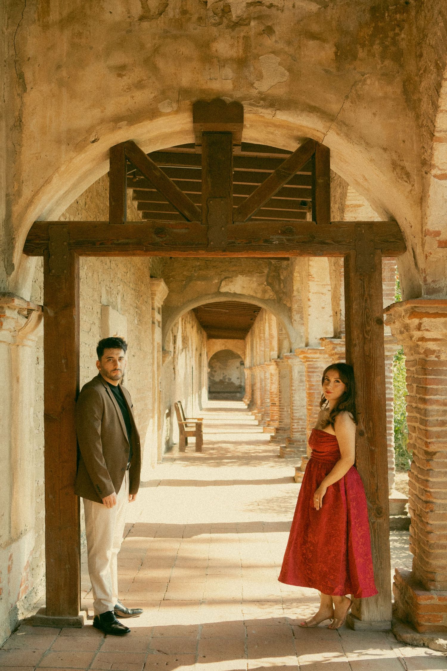 Couple poses in sunlit arched corridor of historic Spanish mission, woman in red dress, man in blazer.
