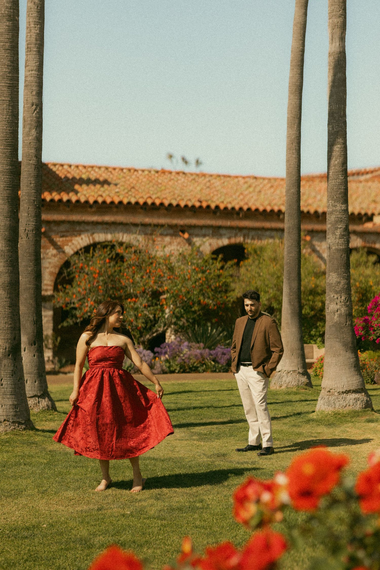 A woman in a red dress and a man in a brown jacket stand on green grass surrounded by palm trees near a Spanish-style building.