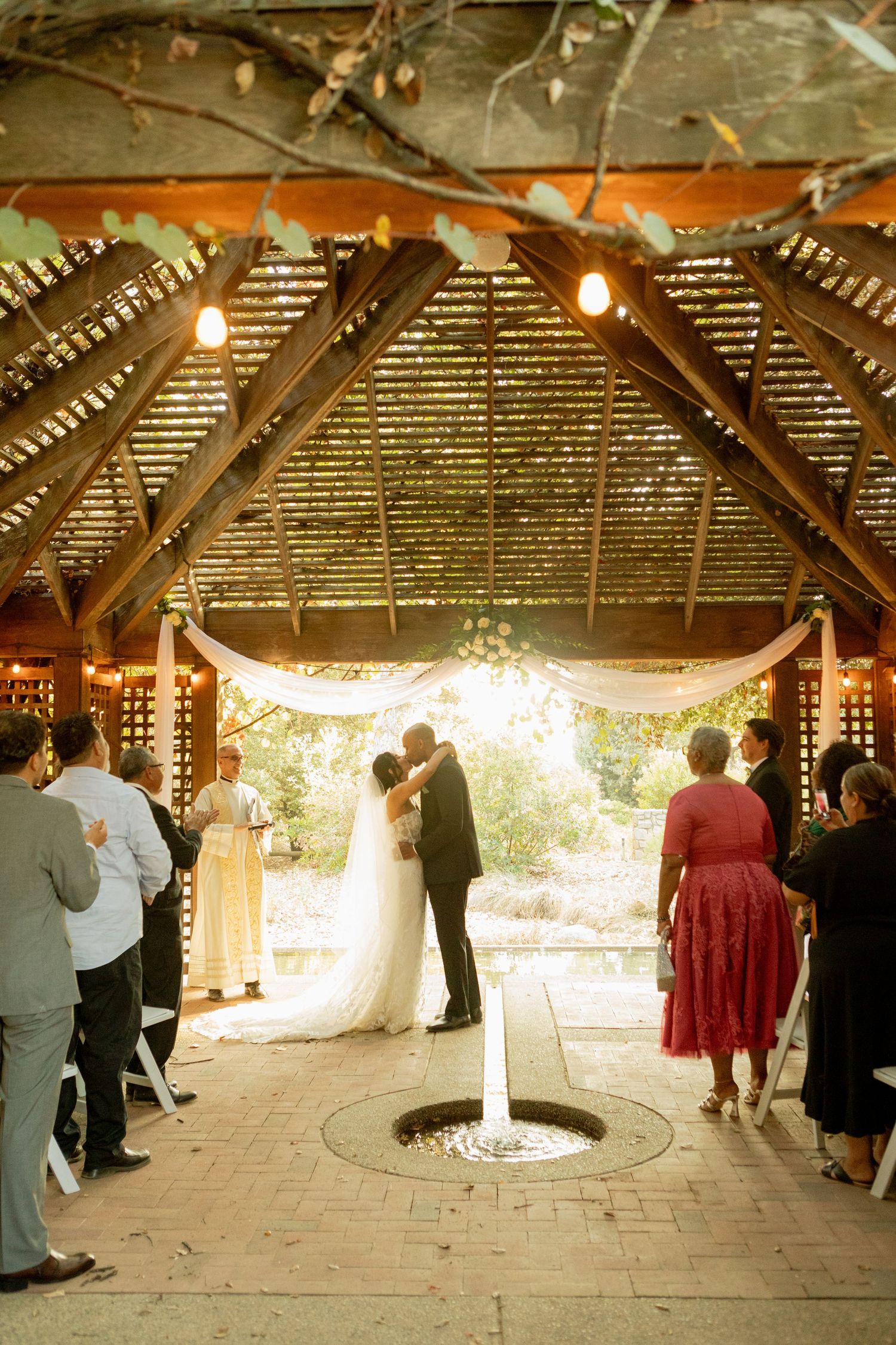 Bride and groom sharing a first kiss under a rustic wooden pergola during their ceremony.