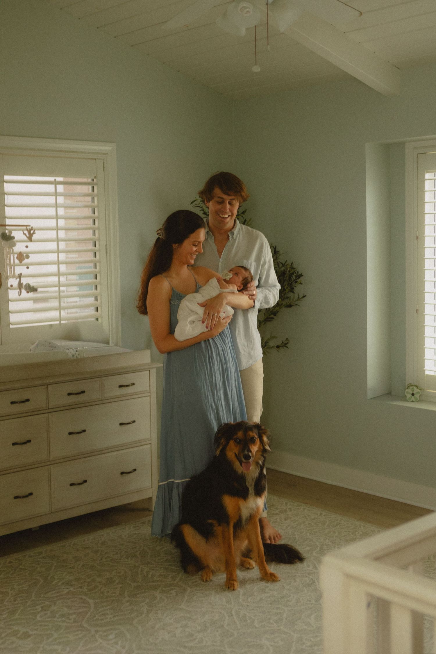 Happy young family with newborn baby and German Shepherd dog standing in nursery room.
