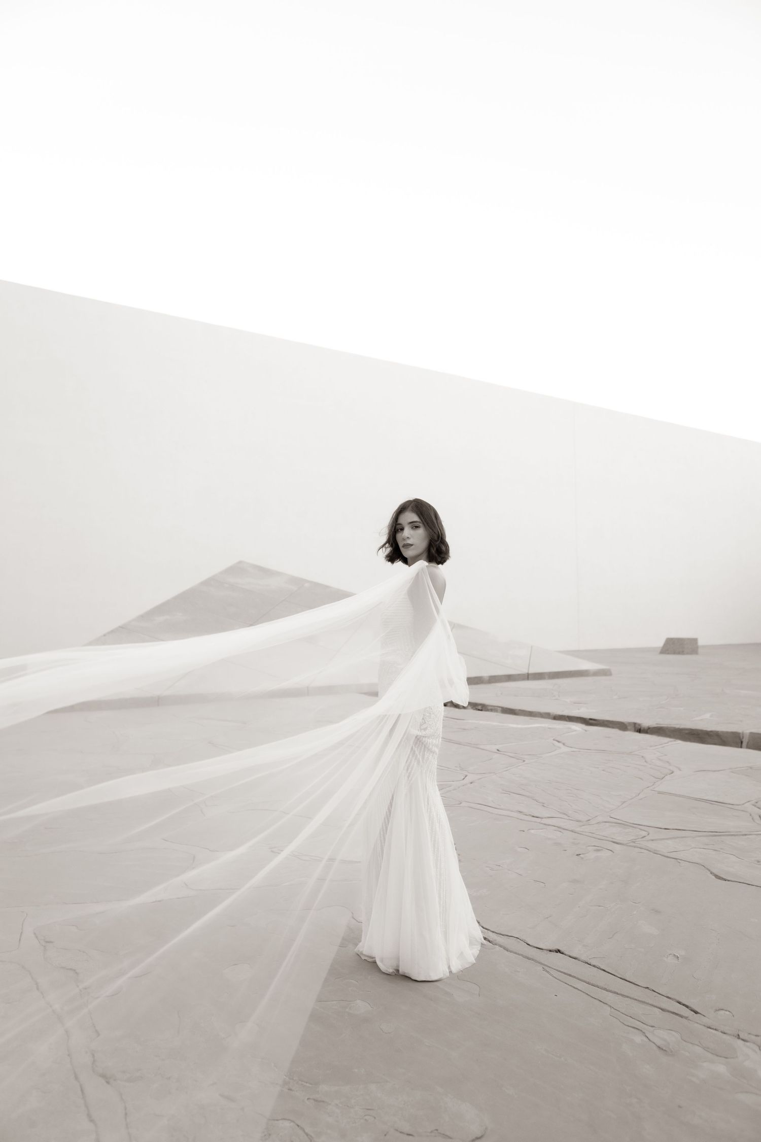 Elegant bride in flowing white gown with dramatic veil billowing in wind against minimalist white backdrop.