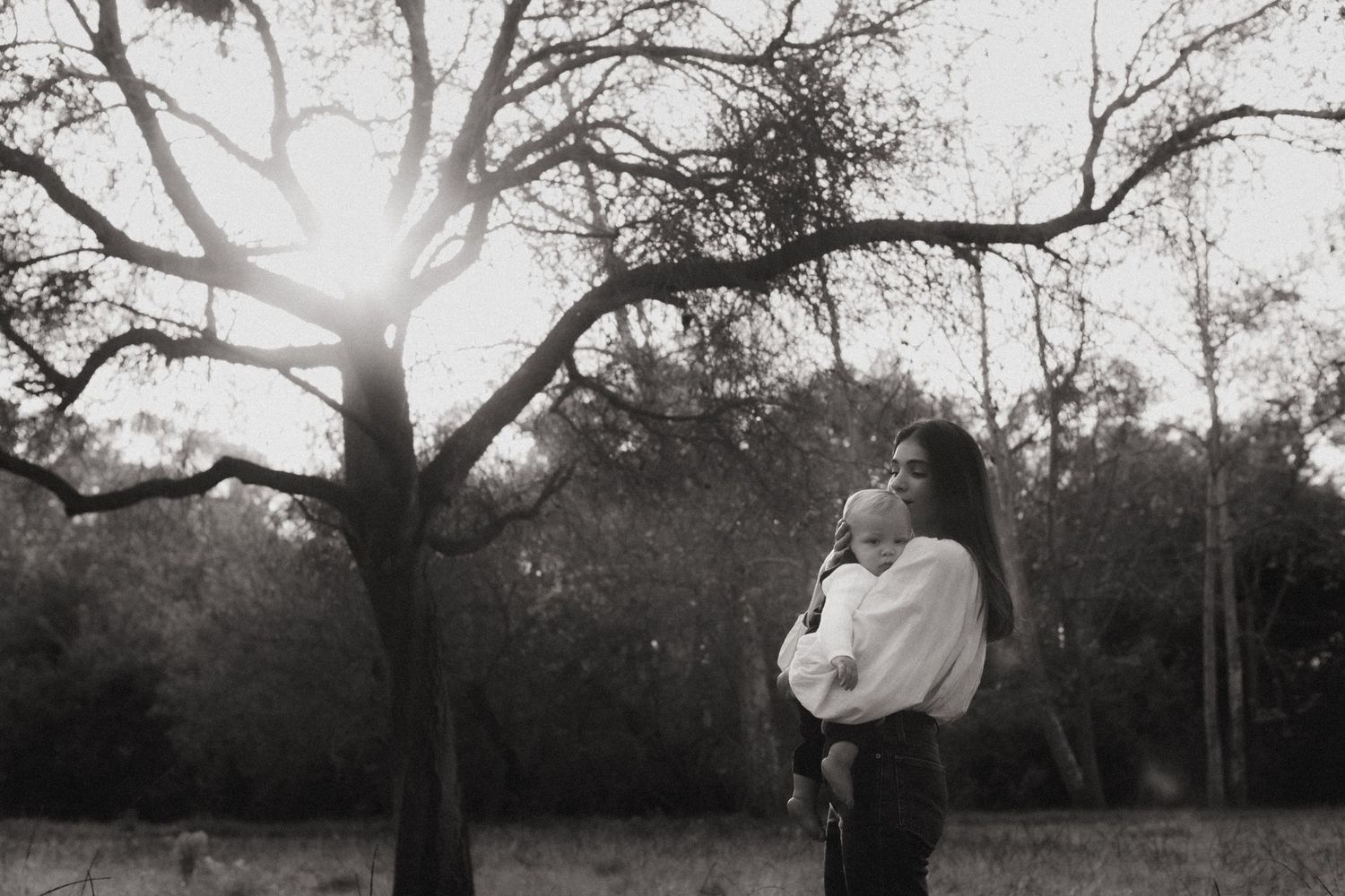 Black and white photo of mother holding newborn baby under bare tree branches in soft backlight.