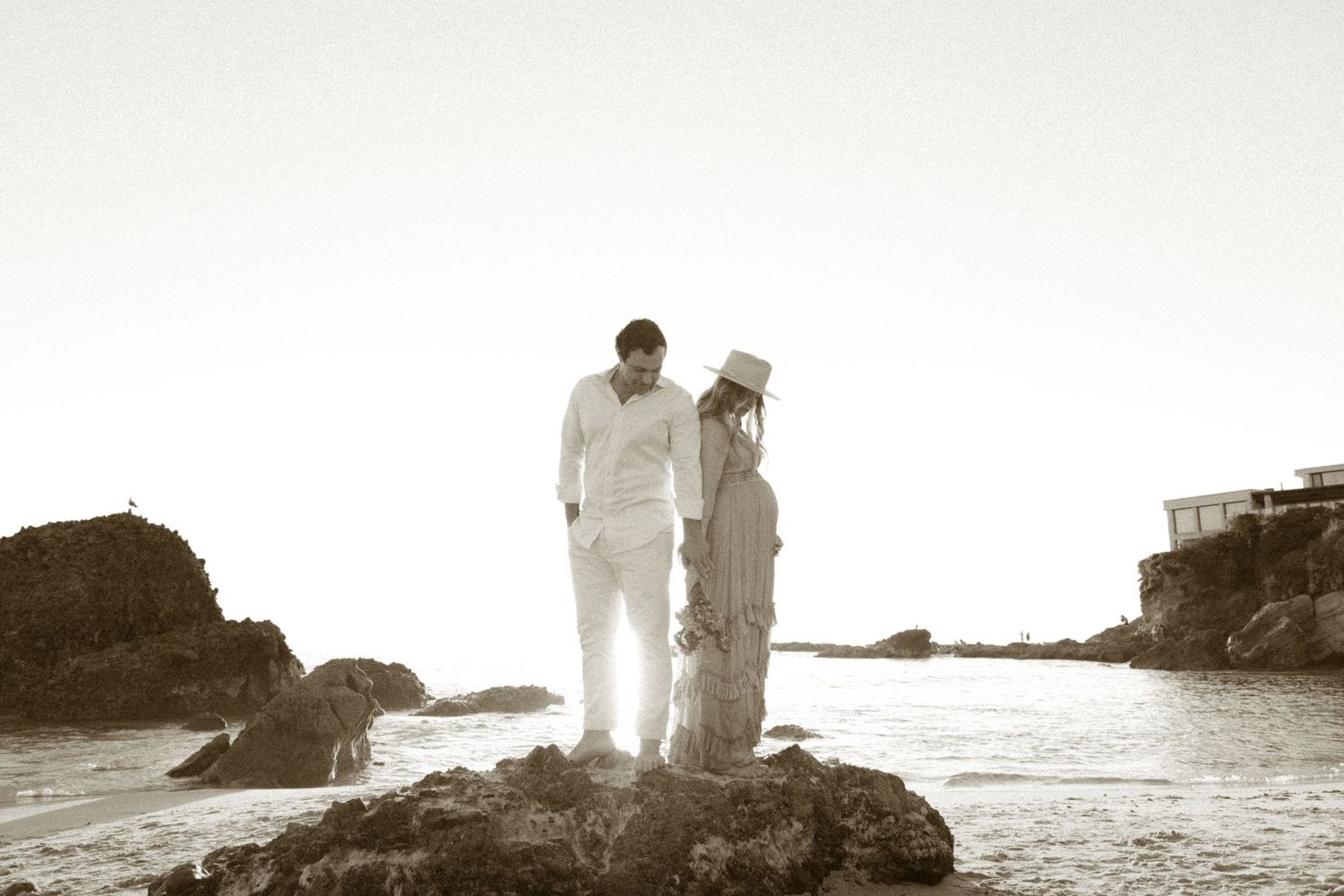 A couple stands on coastal rocks at sunset in a romantic sepia-toned beach portrait.