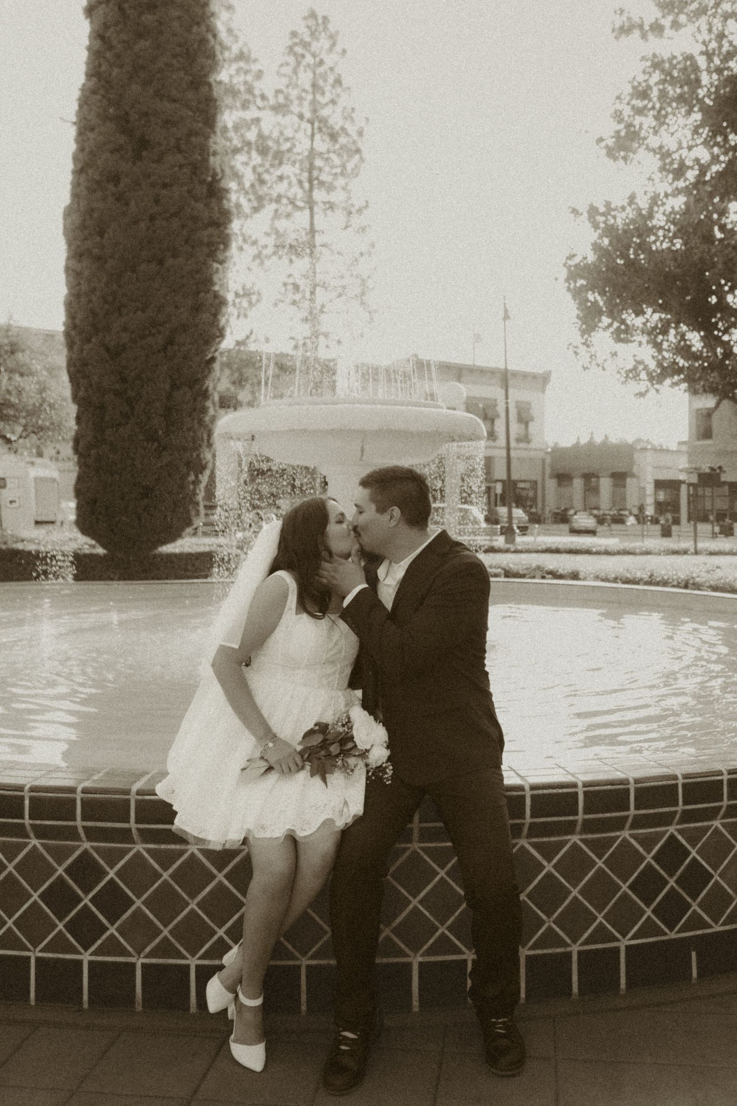 Bride and groom share a romantic kiss by a fountain in a sepia-toned wedding photo outdoors.
