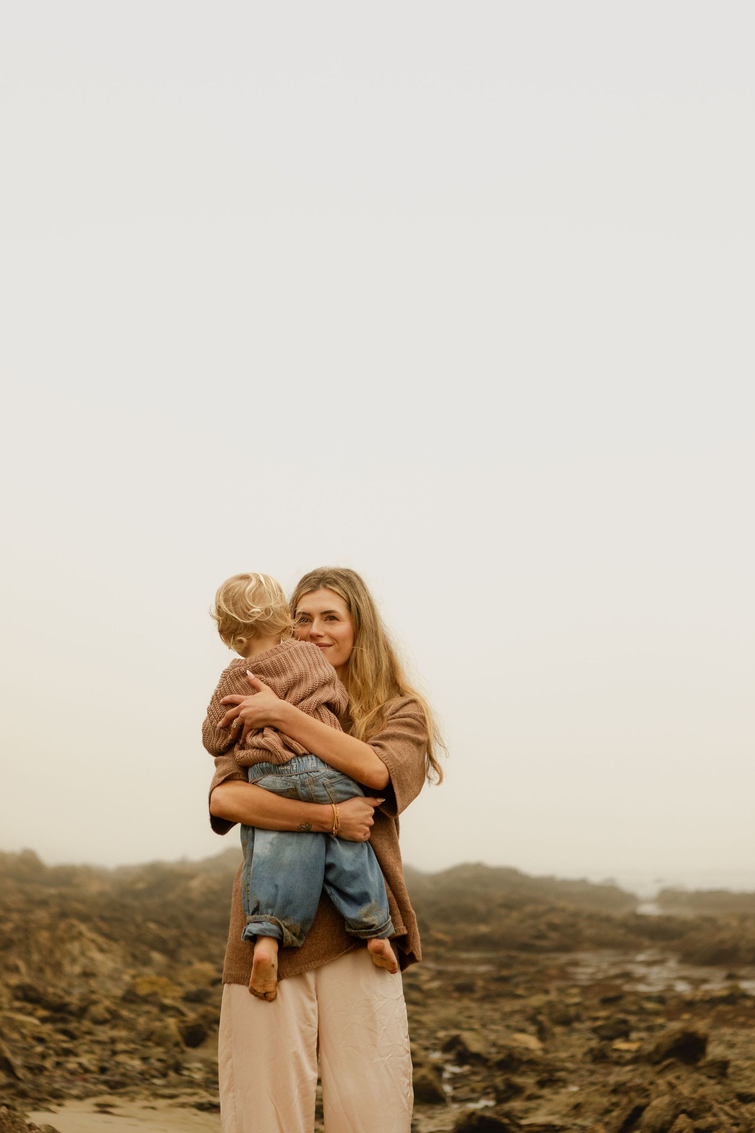 Smiling mother with long blonde hair holding and hugging her toddler child on rocky beach.
