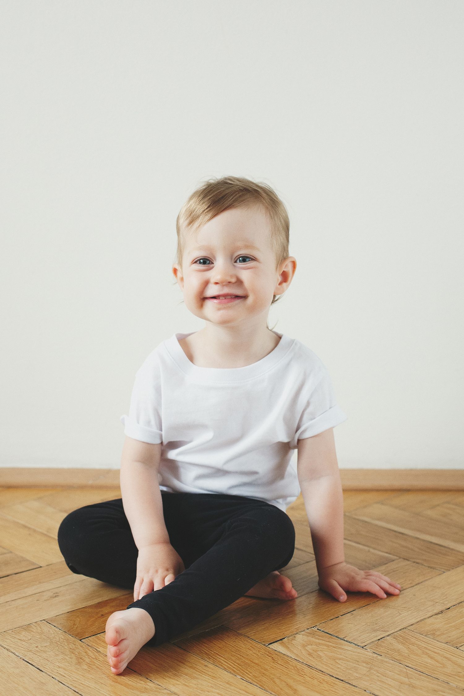 A toddler in white shirt and black pants sits cross-legged on wooden flooring.