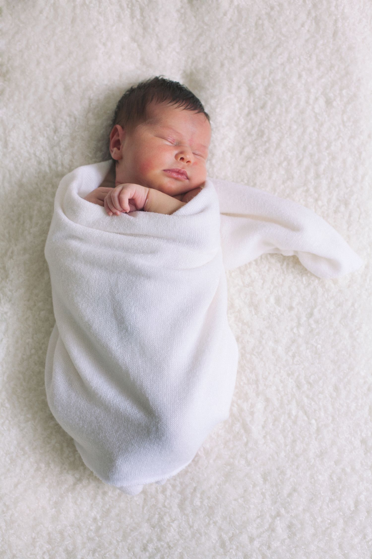 Newborn baby wrapped in soft white blanket against white textured background.