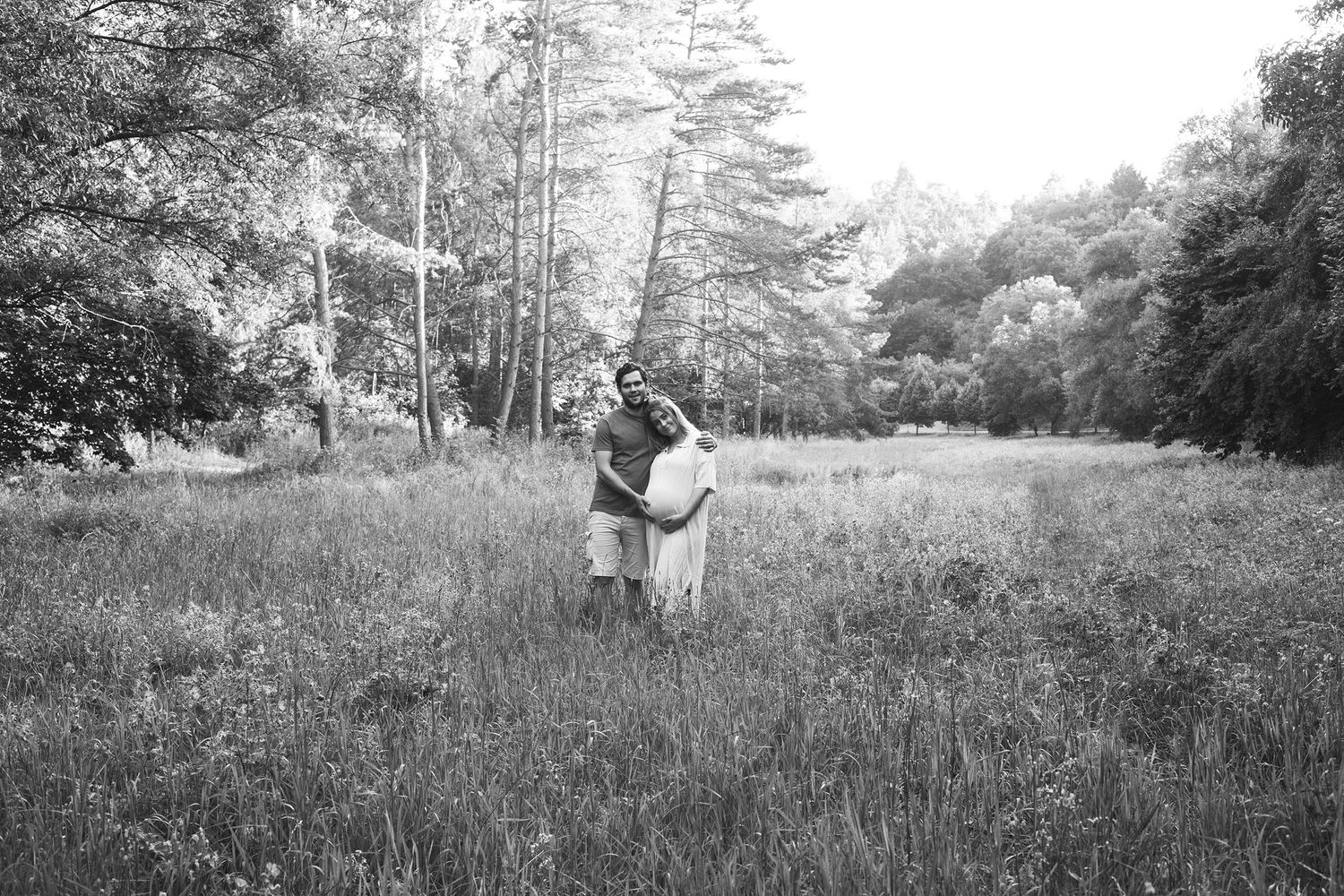 A couple embraces while standing in a serene meadow surrounded by tall trees.