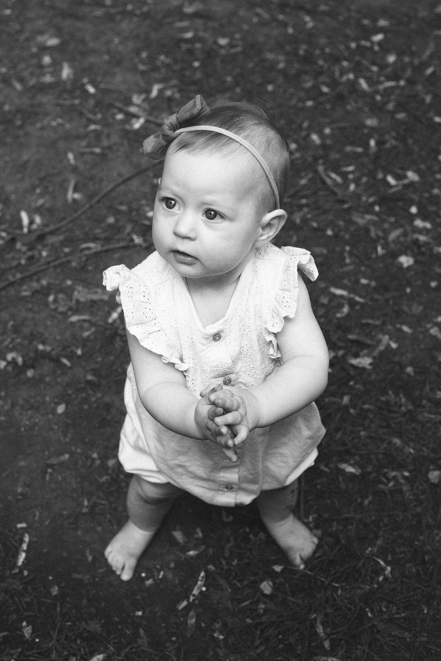 Black and white photos of a baby in a white dress sitting on dark ground.