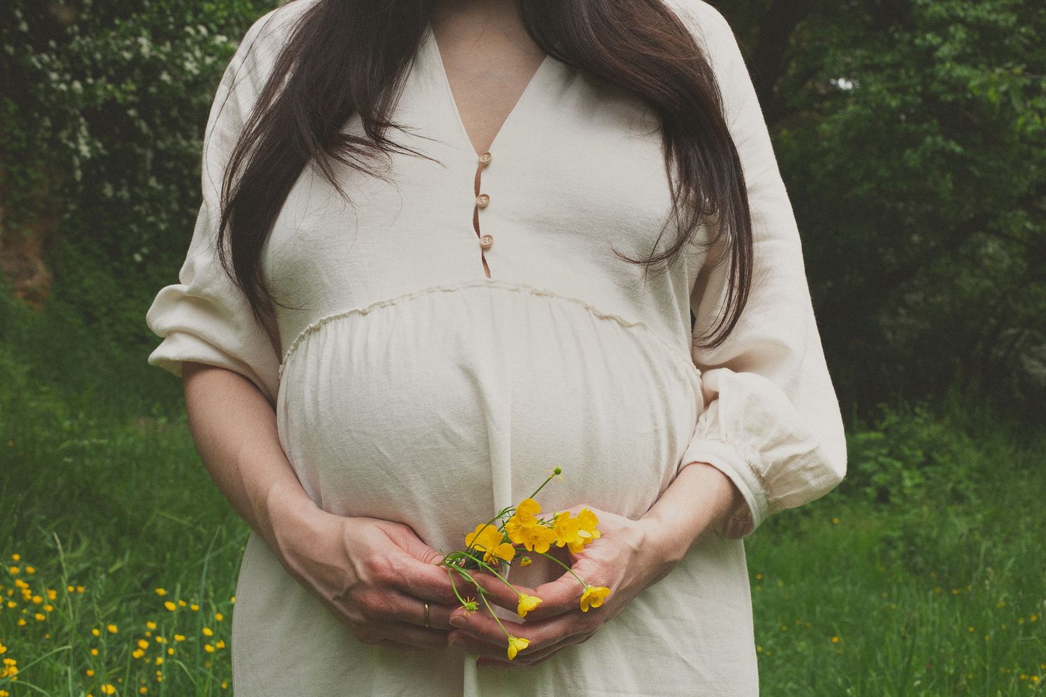 A pregnant figure in a white dress holds yellow wildflowers in a lush green garden setting.