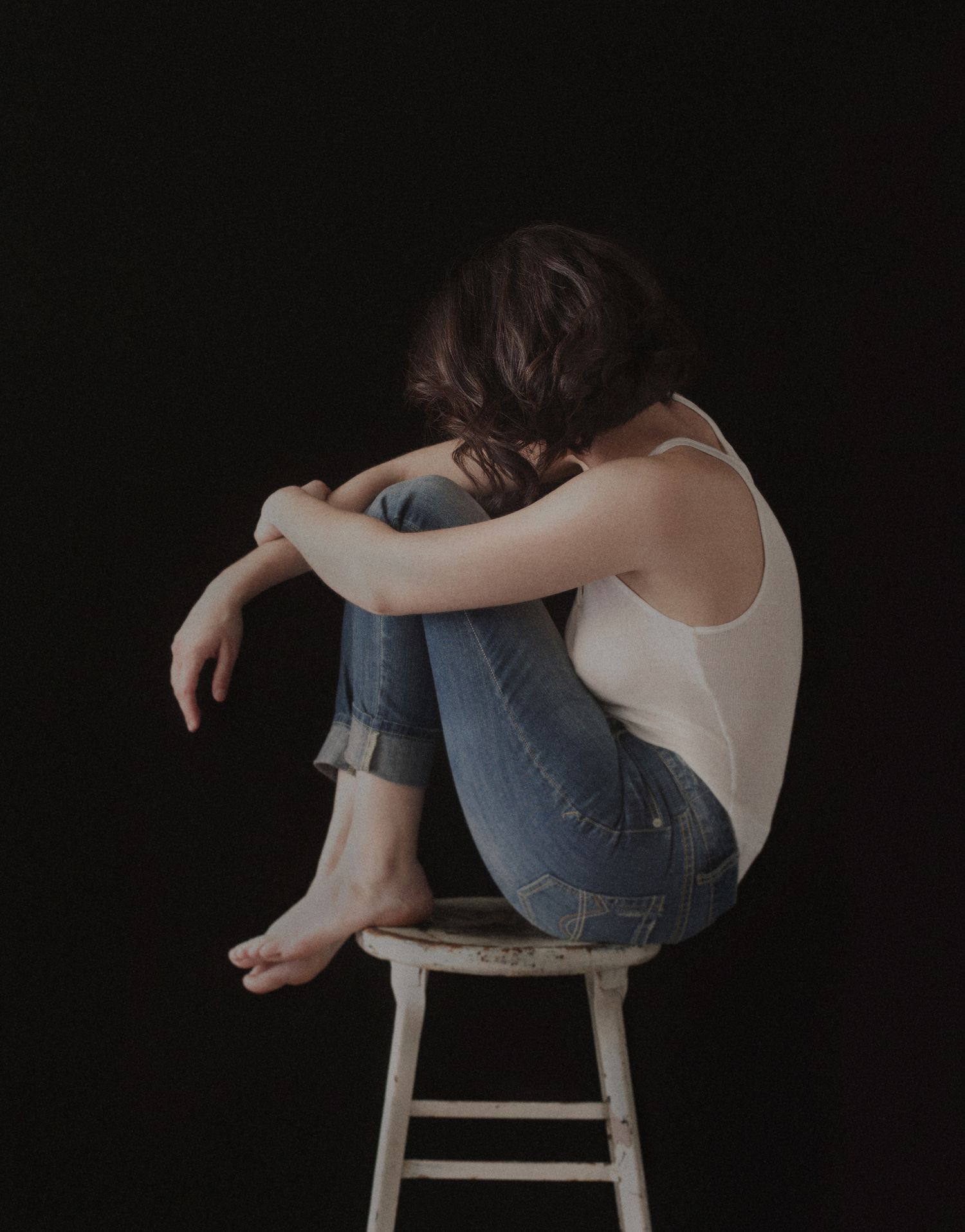 Person in white top and jeans sits curled up on wooden stool against dark background.