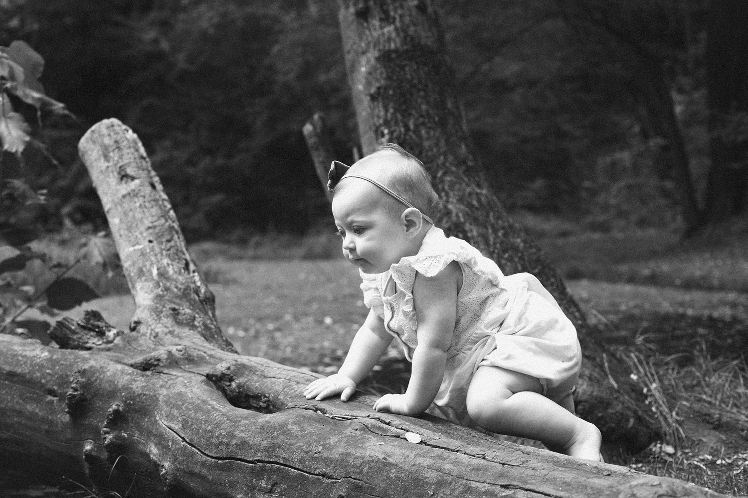 Black and white photo of a child sitting near a tree trunk outdoors.