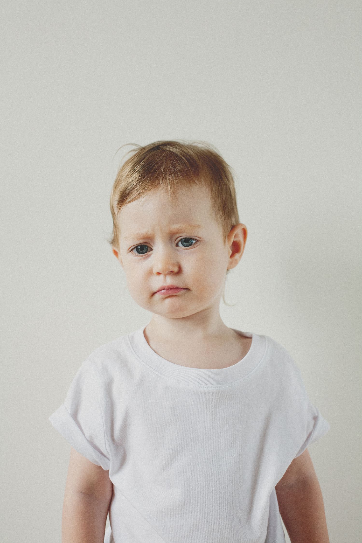 A small child wearing a white shirt poses against a minimalist white backdrop.