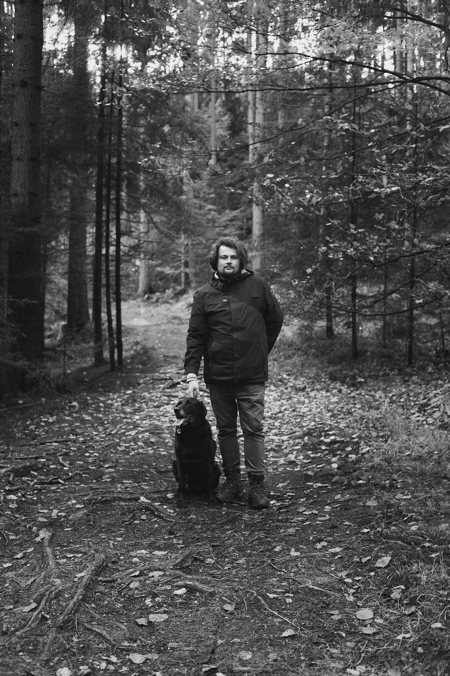Black and white photograph of figures walking through a wooded forest path covered in fallen leaves.