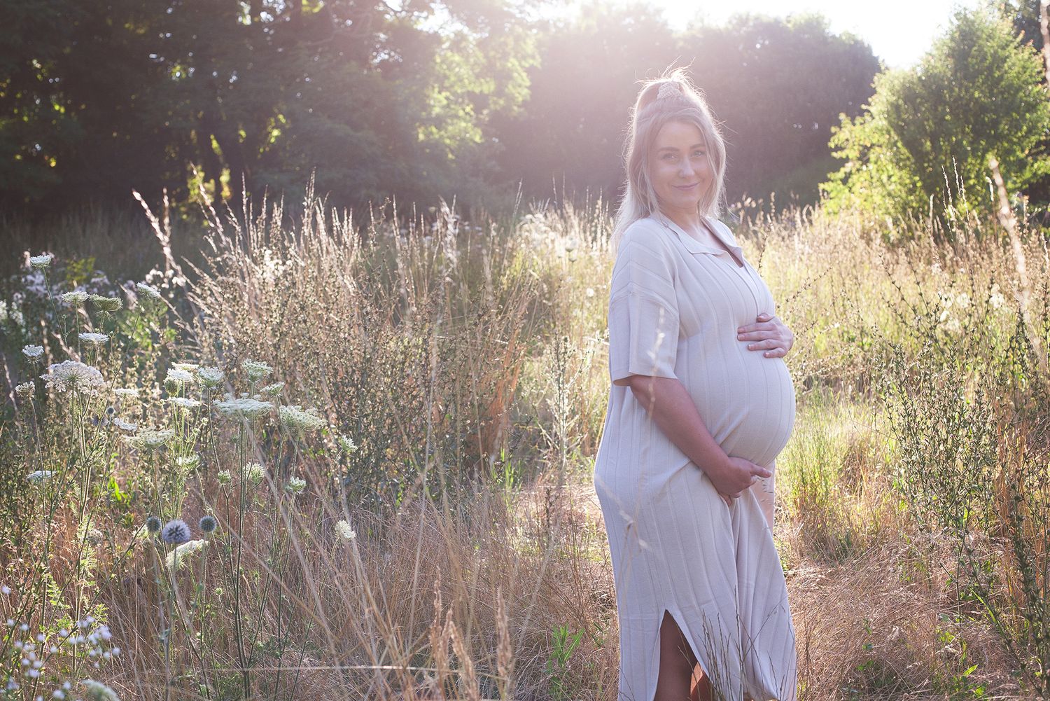 A maternity photo showing a pregnant figure in white dress backlit by golden sunlight in a meadow.