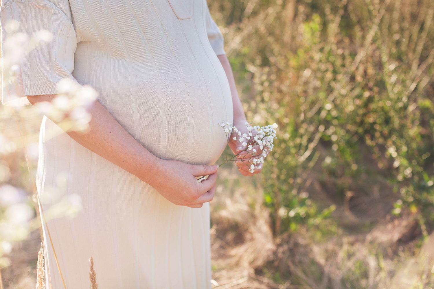 Expecting mother in white dress standing among wildflowers during golden hour.
