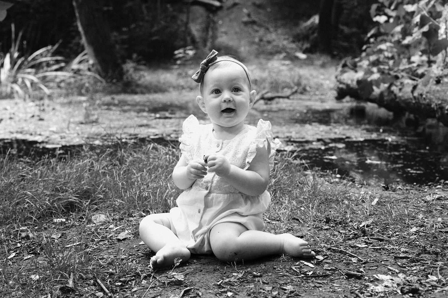 Black and white photo of a small child sitting on grass near a tree trunk.