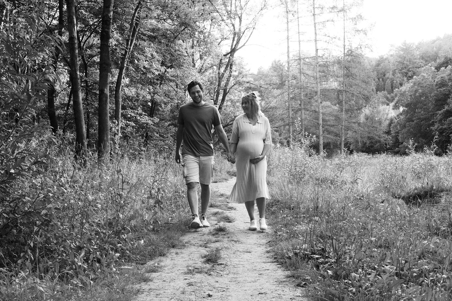 A couple walks hand in hand down a wooded trail during a maternity photoshoot.