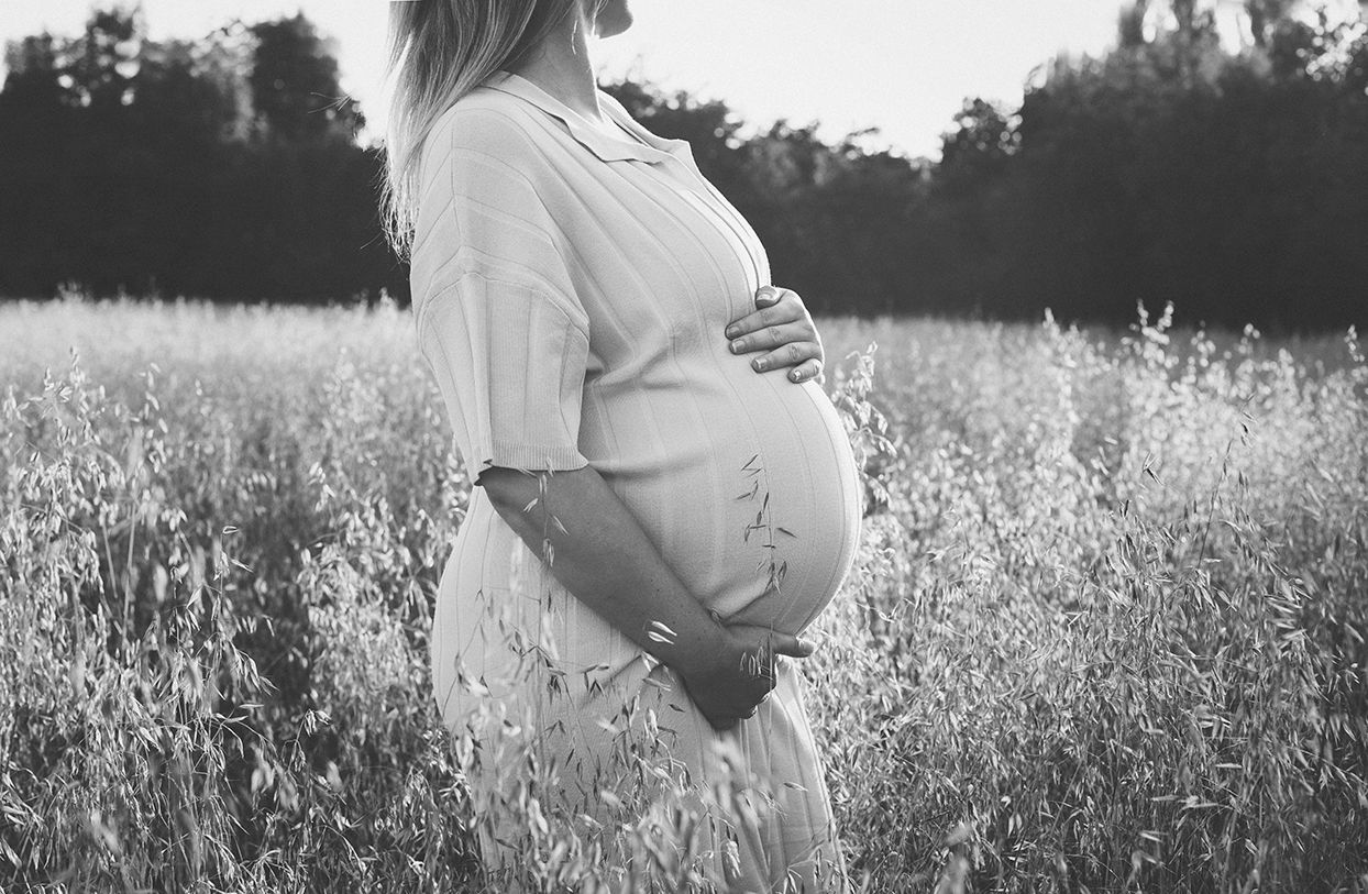 A pregnant woman in a white dress stands in a wildflower meadow during golden hour, captured in black and white.