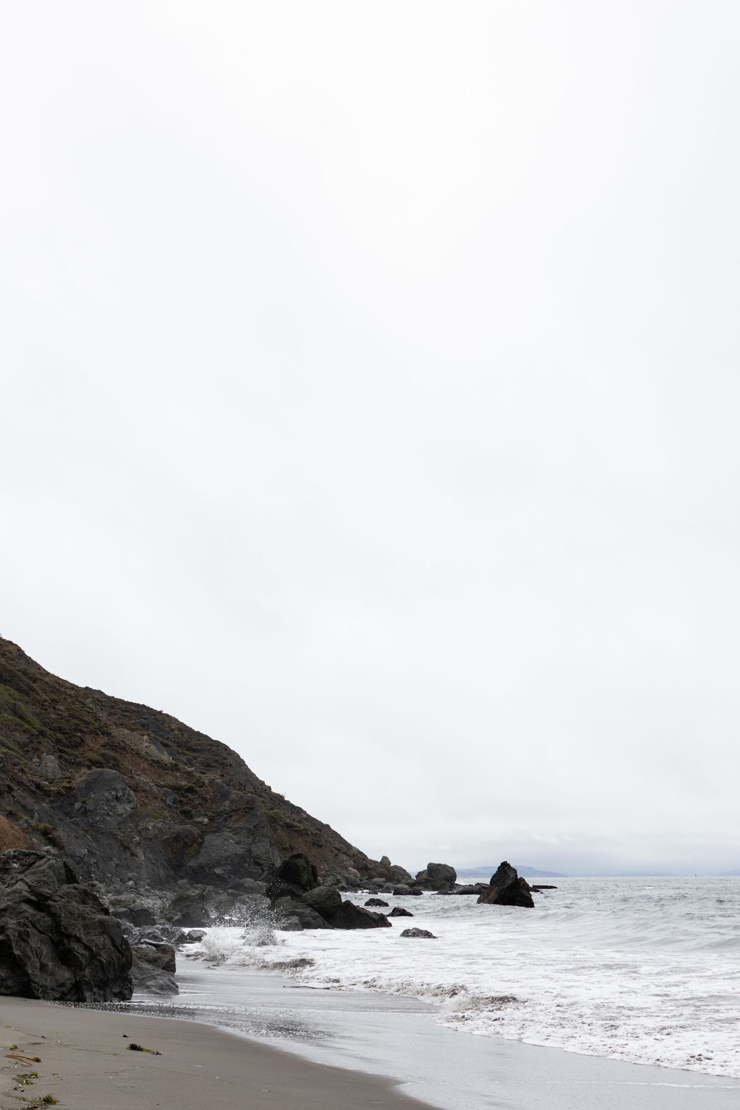 Misty Muir Beach with rocky cliffs, dark boulders, and gentle ocean waves under an overcast sky.