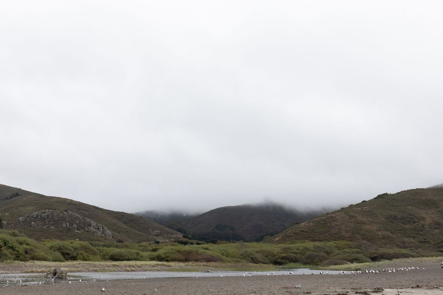Misty mountain landscape with low clouds over rolling hills beside a calm river valley on an overcast day.
