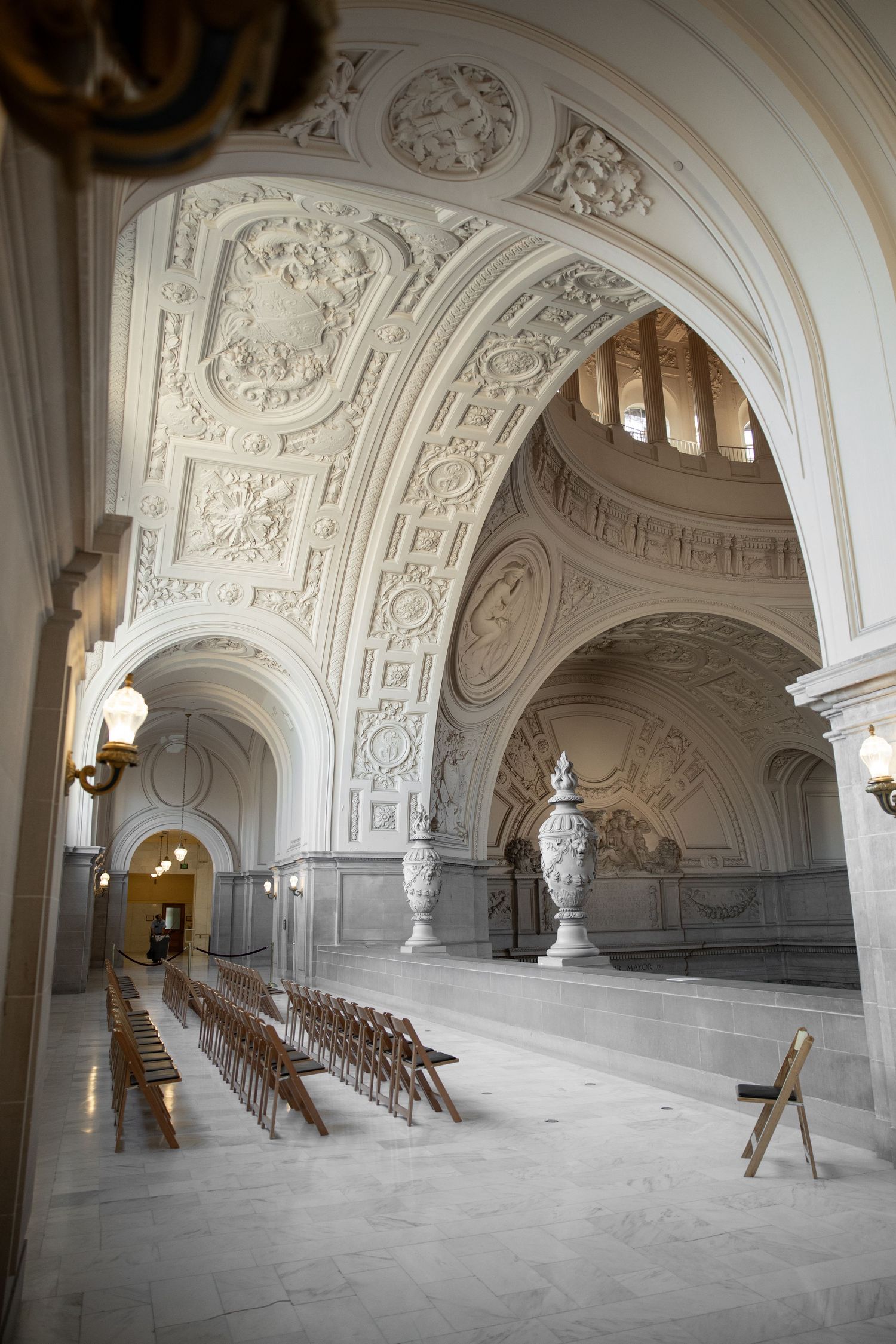 Ornate white marble interior of San Francisco City Hall with arched ceilings, intricate moldings, and rows of wooden chairs.