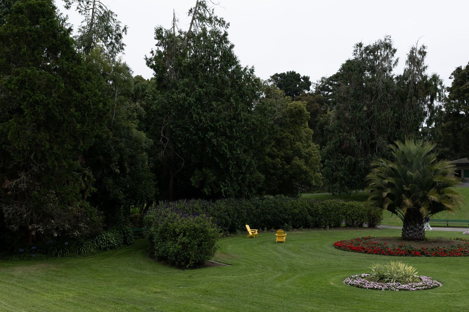 Lush green park garden with manicured lawn, dense trees, colorful flower beds, and yellow chairs on a cloudy day.